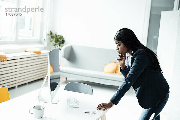 Nachdenkliche Geschäftsfrau mit der Hand am Kinn und Blick auf den Desktop-PC im Büro