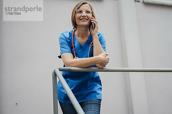 Lächelnder Arzt  der über ein Smartphone spricht und sich an ein Geländer vor der Wand lehnt