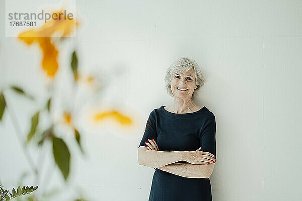 Happy senior woman with arms crossed standing in front of wall