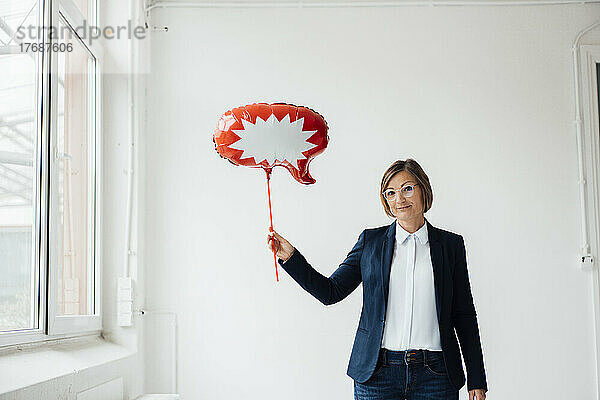 Lächelnde Geschäftsfrau mit Sprechblasenballon vor der Wand im Büro