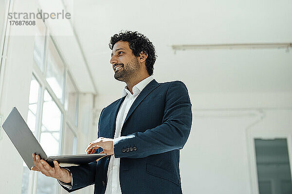 Smiling businessman holding laptop standing in office