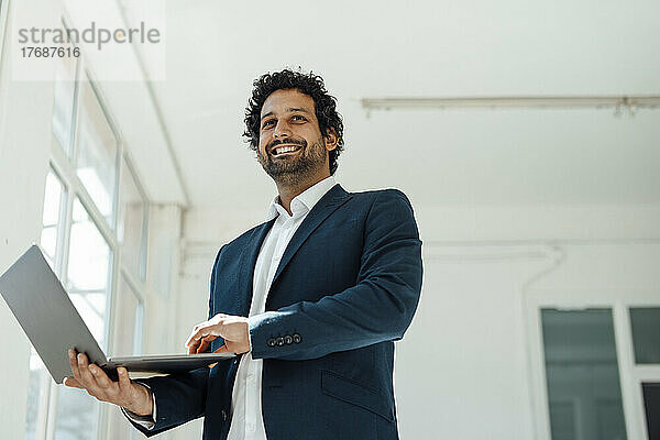 Happy businessman holding laptop standing in office