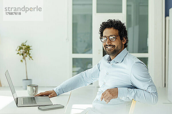 Glücklicher Geschäftsmann mit Laptop und Mobiltelefon sitzt am Schreibtisch im Büro