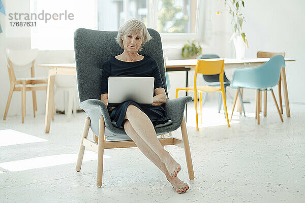 Ältere Geschäftsfrau sitzt im Sessel im Büro und benutzt Laptop