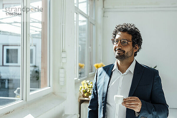 Lächelnder Geschäftsmann mit Kaffeetasse und Blick durch das Fenster im Büro