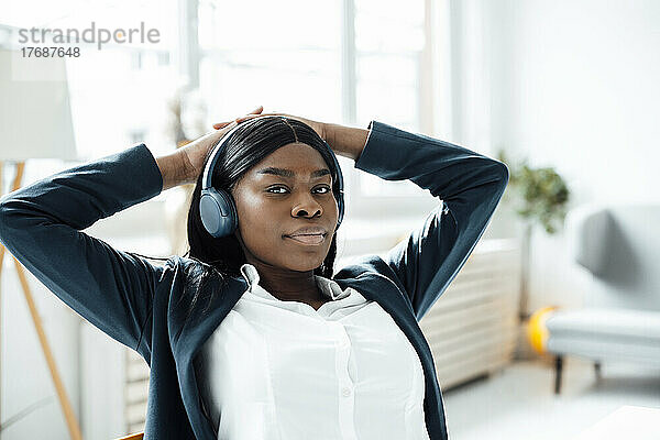 Geschäftsfrau mit Händen hinter dem Kopf  die im Büro Musik über drahtlose Kopfhörer hört