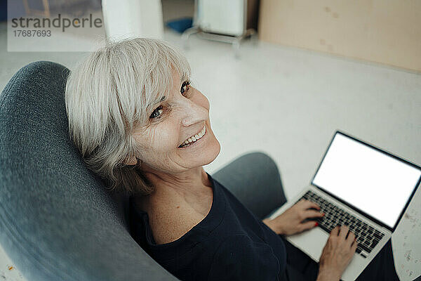 Happy senior businesswoman with laptop sitting in armchair in office
