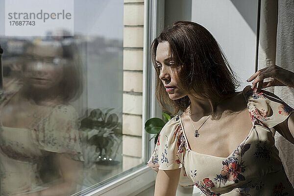 Reife Frau mit braunen Haaren steht zu Hause am Glasfenster