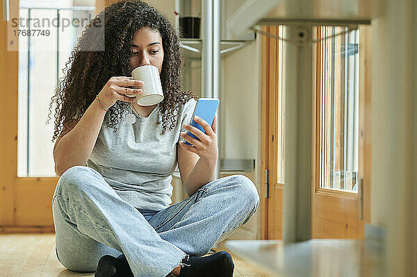 Junge Frau trinkt Kaffee und benutzt ihr Mobiltelefon  während sie zu Hause auf dem Boden sitzt