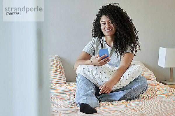 Lächelnde Frau mit Mobiltelefon sitzt zu Hause im Bett