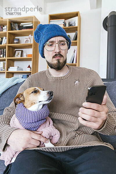 Mann mit Wollmütze sitzt auf Couch und hält Hund in der Hand und checkt Smartphone