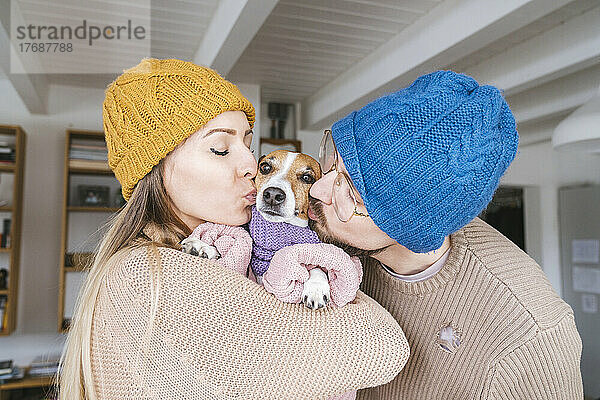 Paar mit Wollmütze zu Hause küsst Hund mit Schal