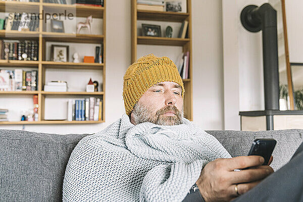 In eine Decke gehüllter Mann mit Wollmütze sitzt auf der Couch und checkt sein Smartphone