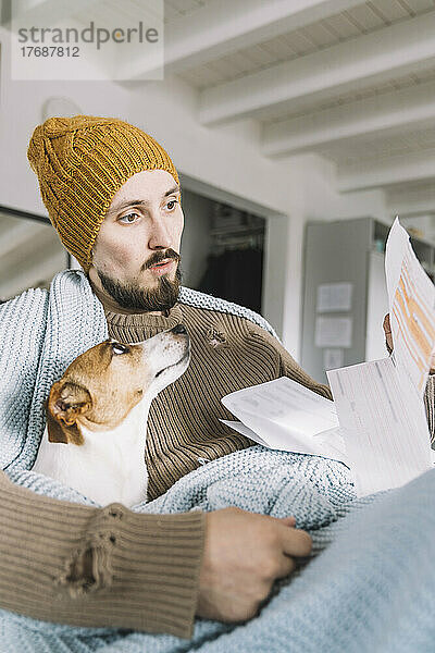 Wickede  NRW  Deutschland. Ein junger Mann trägt eine Strickmütze Zuhause mit einem Hund Jack Russell Terrier schaut erstaunlich auf eine Rechnung an
