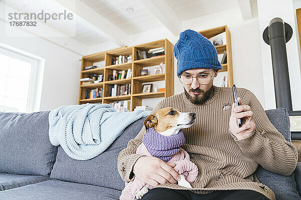 Mann mit Wollmütze sitzt auf Couch und hält Hund in der Hand und checkt Smartphone