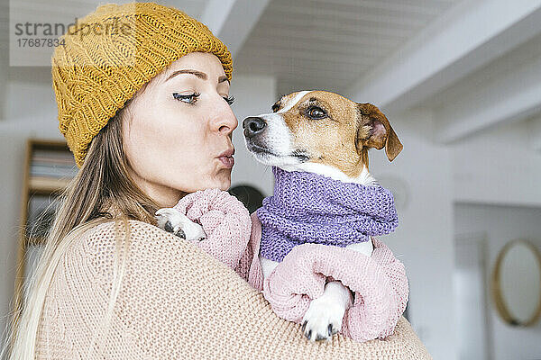 Woman wearing wooly hat at home kissng dog with scarf