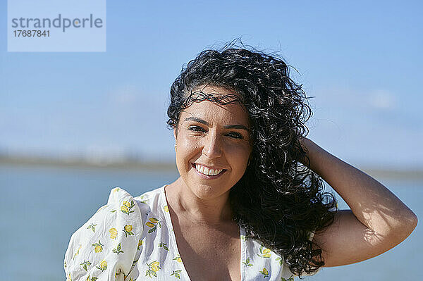 Glückliche junge Frau mit der Hand im Haar am Strand an einem sonnigen Tag