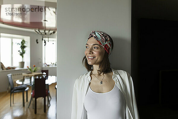 Lächelnde reife Frau mit Stirnband lehnt zu Hause an der Wand