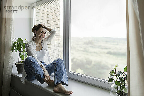 Glückliche reife Frau mit der Hand im Haar sitzt zu Hause auf der Fensterbank