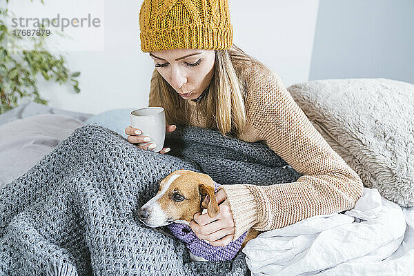 Junge Frau mit Wollmütze trinkt heißen Tee und sitzt mit Hund im Bett