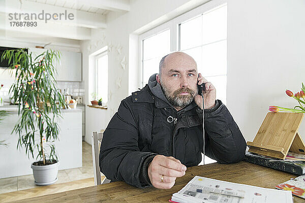 Reifer Mann sitzt zu Hause in Winterjacke und telefoniert