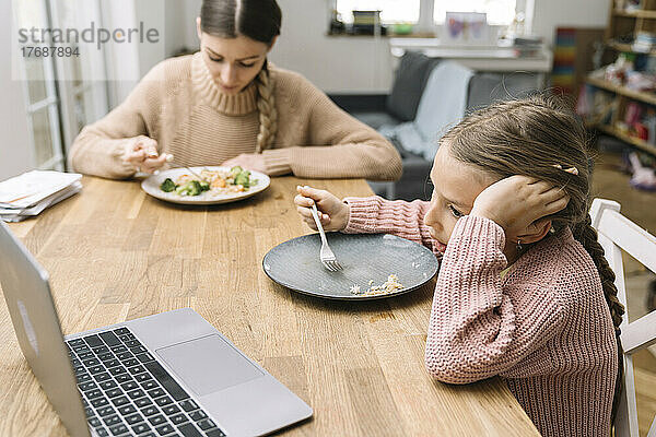 Traurige junge Frau und gelangweiltes Mädchen essen zu Hause und schauen auf den Laptop