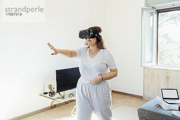 Junge Frau mit Virtual-Reality-Simulator gestikuliert im heimischen Wohnzimmer