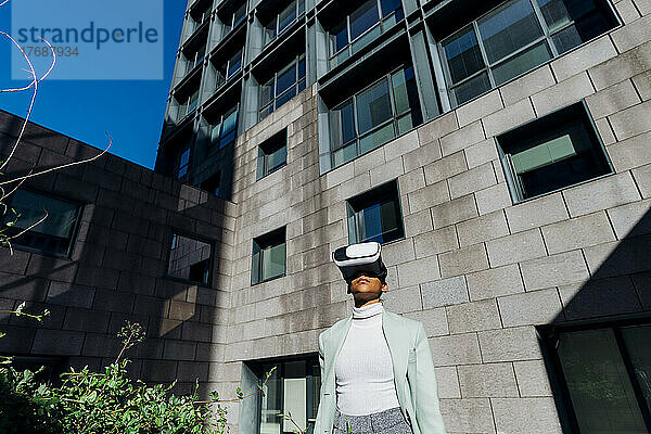 Junge Geschäftsfrau mit VR-Brille vor Bürogebäude an sonnigem Tag
