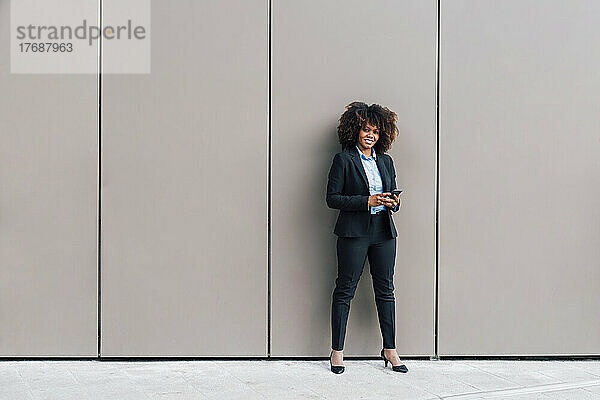 Lächelnde Geschäftsfrau mit Mobiltelefon vor der Wand