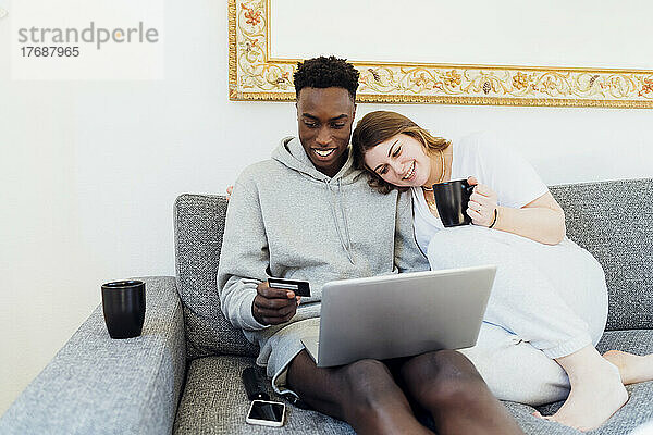 Glückliches junges Paar mit Kreditkarte und Laptop  das zu Hause zusammen auf dem Sofa sitzt