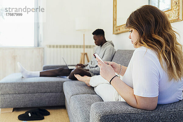 Junge Frau benutzt Smartphone und sitzt mit ihrem Freund zu Hause auf dem Sofa