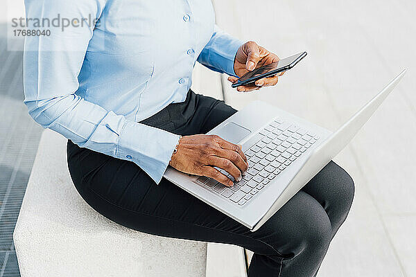 Geschäftsfrau benutzt Mobiltelefon und sitzt mit Laptop auf Betonblock
