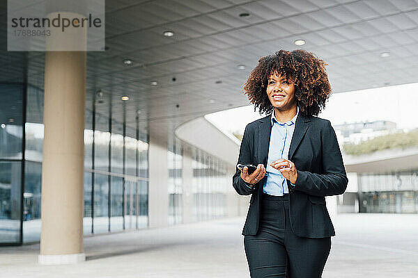 Lächelnde Geschäftsfrau mit Smartphone geht vor Bürogebäude