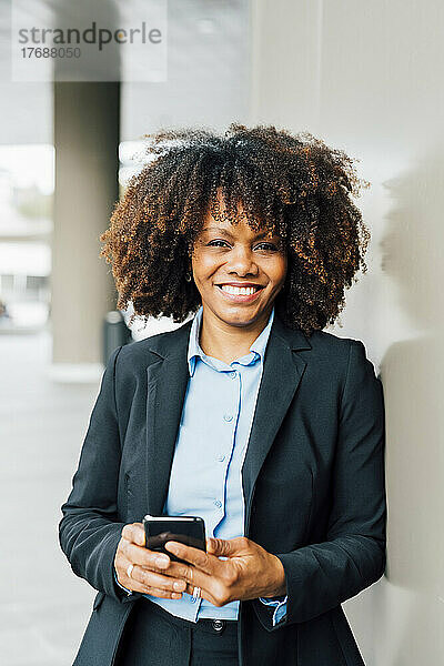 Glückliche Afro-Geschäftsfrau hält ihr Mobiltelefon an die Wand gelehnt