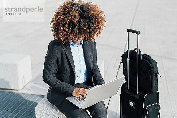 Afro Businesswoman using laptop sitting on concrete block by luggage