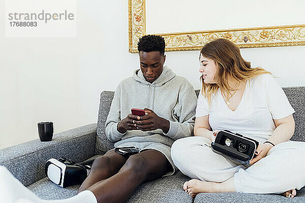 Junger Mann benutzt Smartphone und sitzt zu Hause neben seiner Freundin auf dem Sofa