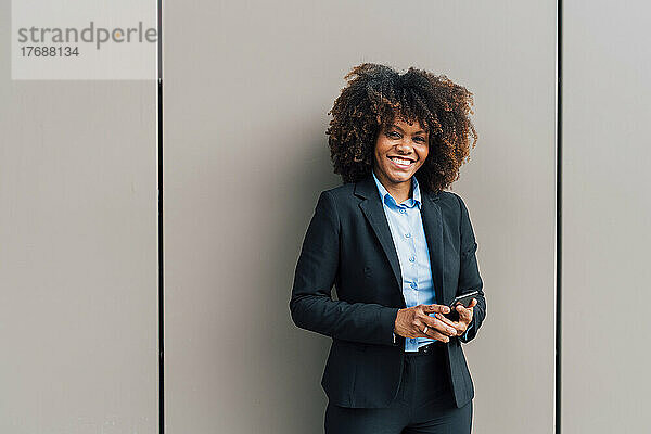 Glückliche Afro-Geschäftsfrau mit Mobiltelefon vor der Wand
