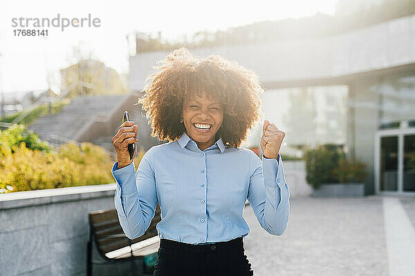 Fröhliche Afro-Geschäftsfrau mit Mobiltelefon feiert an einem sonnigen Tag