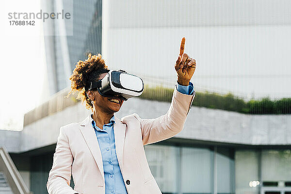 Glückliche Geschäftsfrau mit Virtual-Reality-Simulator gestikuliert vor Bürogebäude