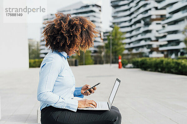 Afro businesswoman using mobile phone sitting with laptop in city