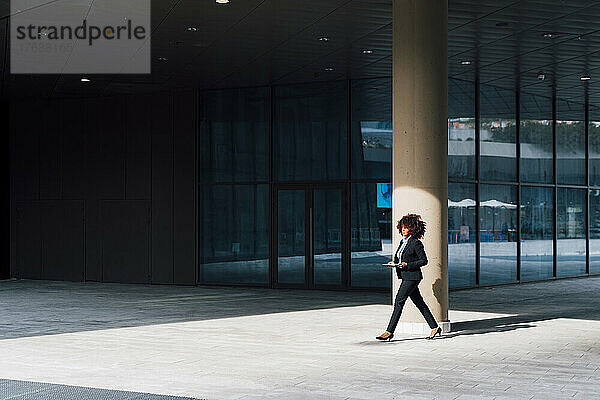 Geschäftsfrau hält Tablet-PC in der Hand und läuft an einem sonnigen Tag vor dem Bürogebäude