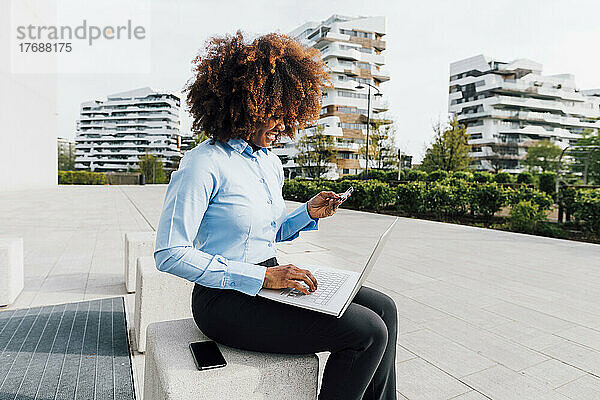Lächelnde Geschäftsfrau mit Kreditkarte beim Online-Einkauf per Laptop auf Betonblock sitzend