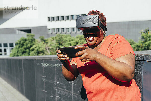 Glückliche Frau  die an einem sonnigen Tag einen Virtual-Reality-Simulator trägt und ihr Smartphone nutzt