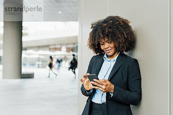 Lächelnde Afro-Geschäftsfrau benutzt Smartphone und lehnt an der Wand