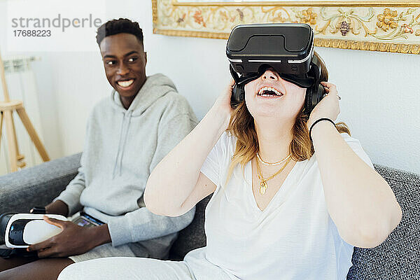 Glückliche Frau mit Virtual-Reality-Simulator sitzt mit ihrem Freund zu Hause auf dem Sofa