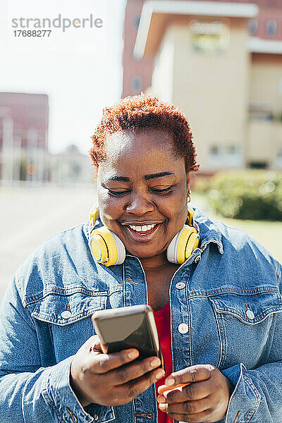 Glückliche Frau mit kabellosen Kopfhörern  die ihr Smartphone auf der Straße benutzt
