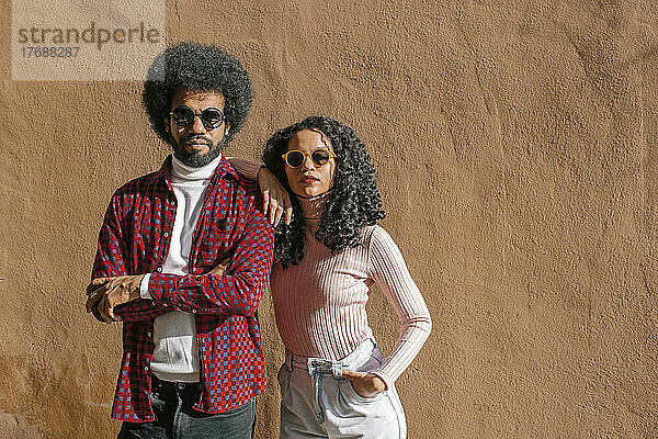 Ein Paar mit Sonnenbrille steht an einem sonnigen Tag vor einer braunen Wand