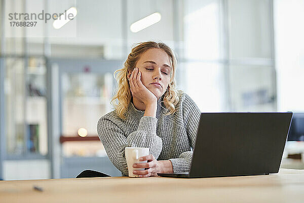 Müde junge Frau sitzt mit Laptop am Schreibtisch im Büro