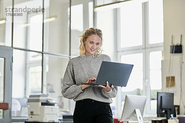 Lächelnde junge Frau steht mit Laptop im Büro