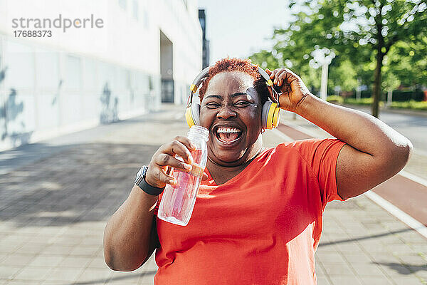 Fröhliche Frau hält eine Wasserflasche in der Hand und hört Musik über kabellose Kopfhörer